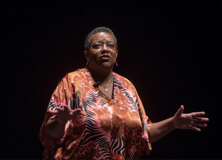 Image is a photograph of performer Sheila Arnold dressed in a printed orange frock against a black background, posed in mid performance.
