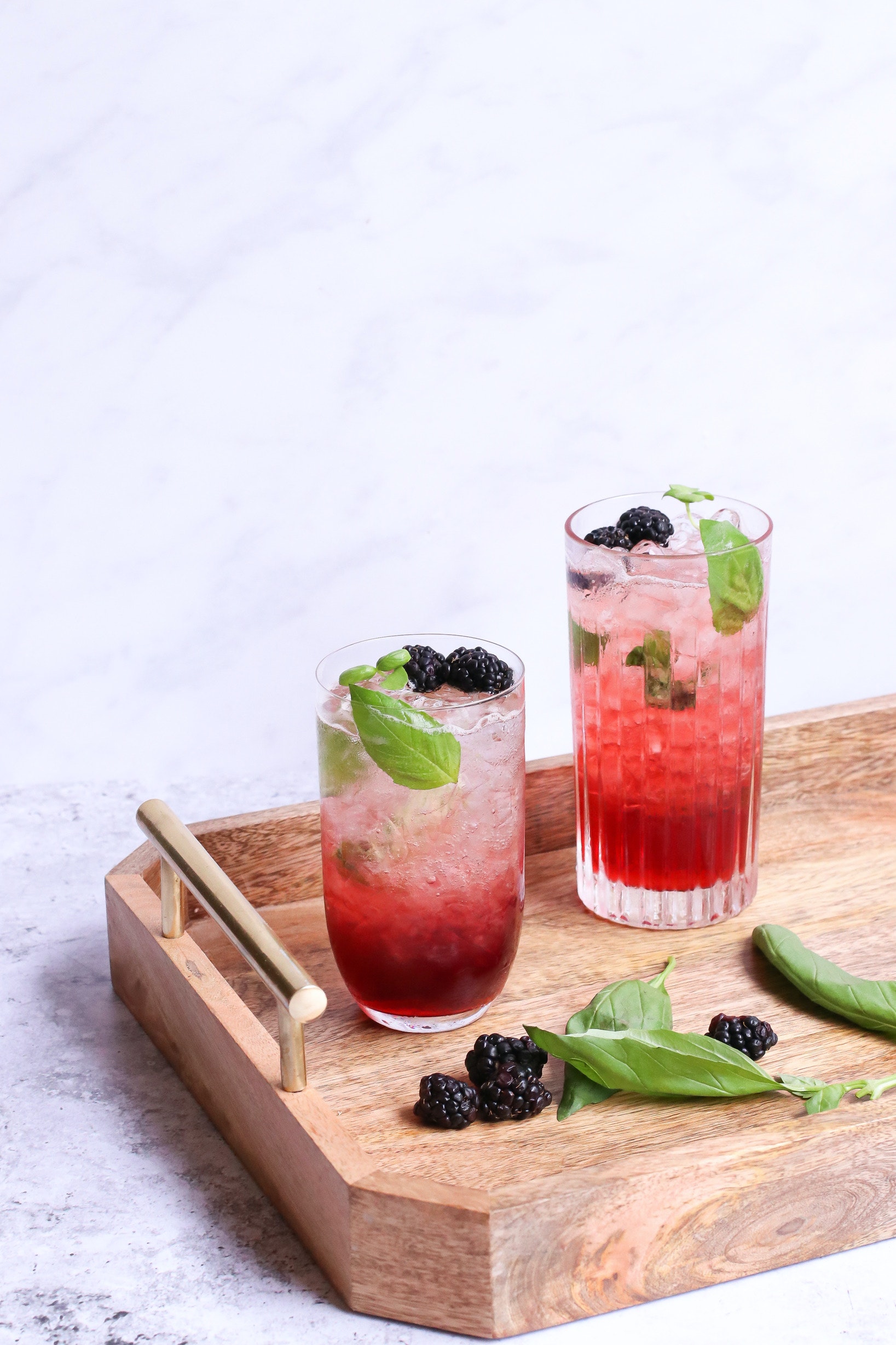 two glasses of red liquid topped with berries and garnishes sit on a fancy wooden tray