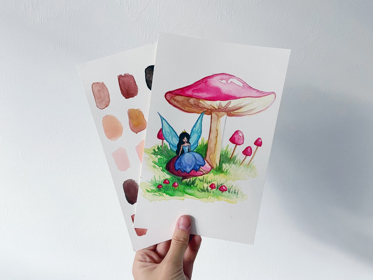 person holds up a watercolor painting of a fairy sitting on a mushroom