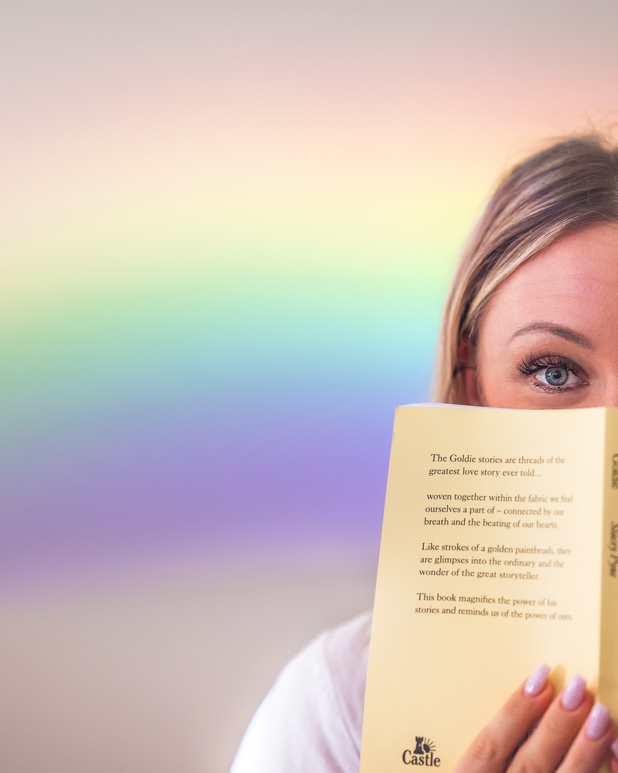 A woman holds a book up in front of her face with a rainbow behind her