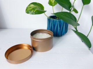 A gold tin of sugar scrub sits on a white shelf in front of an ivy plant