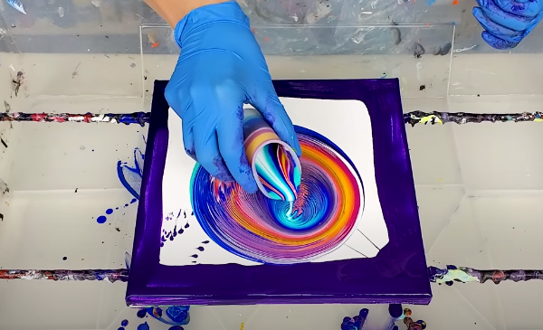 Hand with cup pouring different colors of paint onto a canvas.
