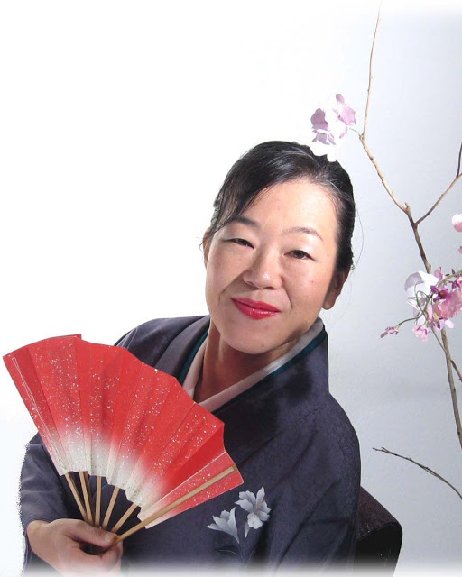 Photo of artist, Kuniko Yamamoto wearing a black robe and holding a red fan.