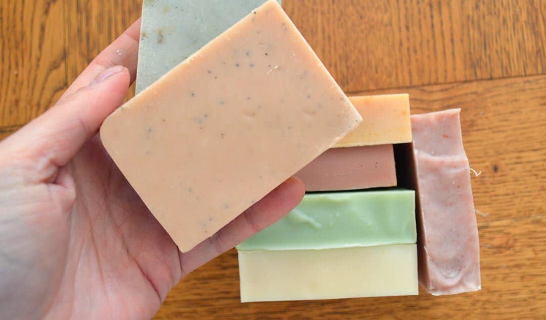 Hand holding a bar of homemade soap with other soap bars in the background.