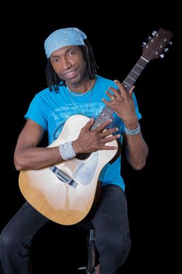 Photo of performer Karlus Trapp wearing a blue t-shirt, bandanna and holding a guitar.
