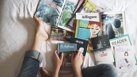 Person in bed with a pile of books and magazines at their feet.