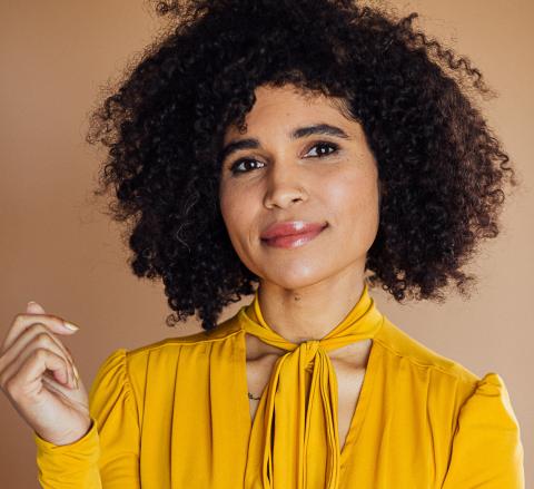 A headshot of Kirya Traber smiling in front of a tan background and wearing a yellow blouse.