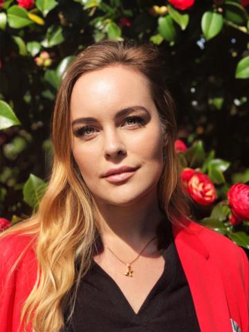 A headshot of writer Rebecca Renner wearing a black blouse and red jacket, in front of green foliage.