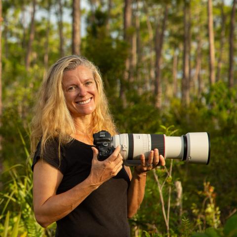 Kirsten Hines hold a white camera with a telephoto lens in the woods.