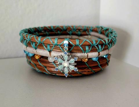 A pine needle basket woven with teal thread and teal and tan beads with a jeweled charm hanging from the rim.