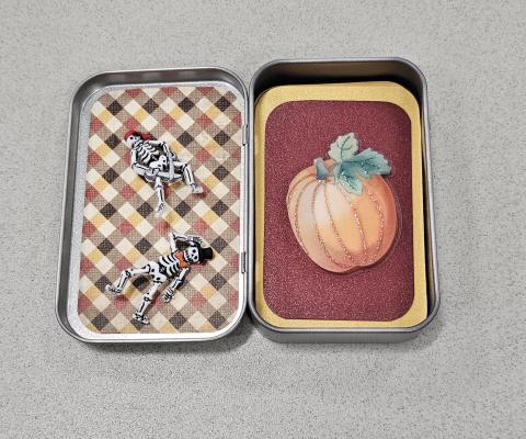 An open Altoid tin with cartoon skeletons on one side and a paper cutout of a pumpkin on the other.