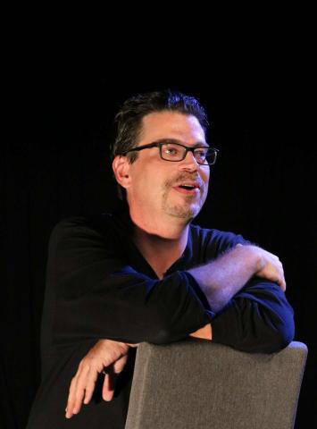 Playwright Glenn Schudel sits on a backward facing chair.