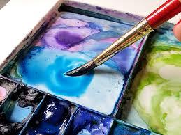 A watercolor paint pan in blue, purple, and green, with a red brush picking up some blue paint.