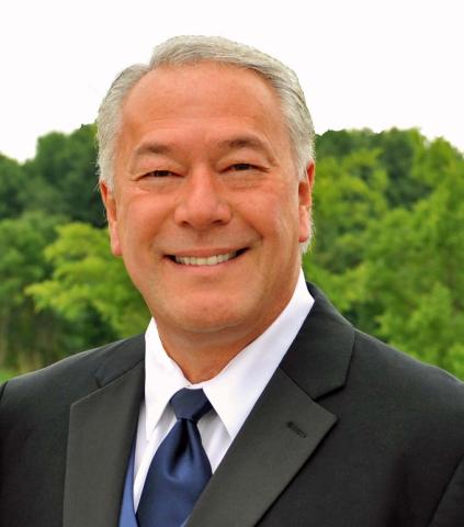 A headshot of a smiling gray haired man wearing a suit jacket and tie in front of green shrubs.