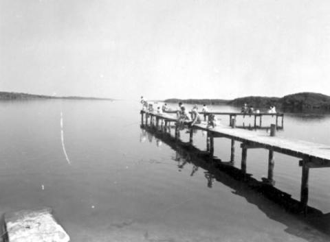 A black and white photograph of people fishing off a pier on a river.