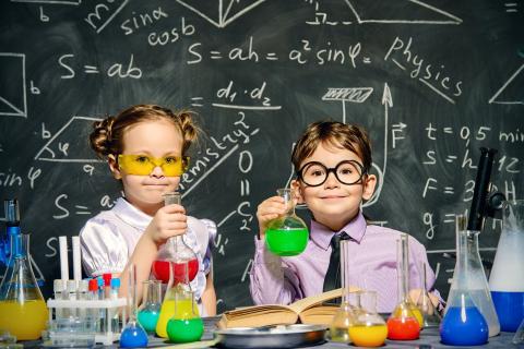 Two children dressed as scientists performing experiments in front of a chalkboard.
