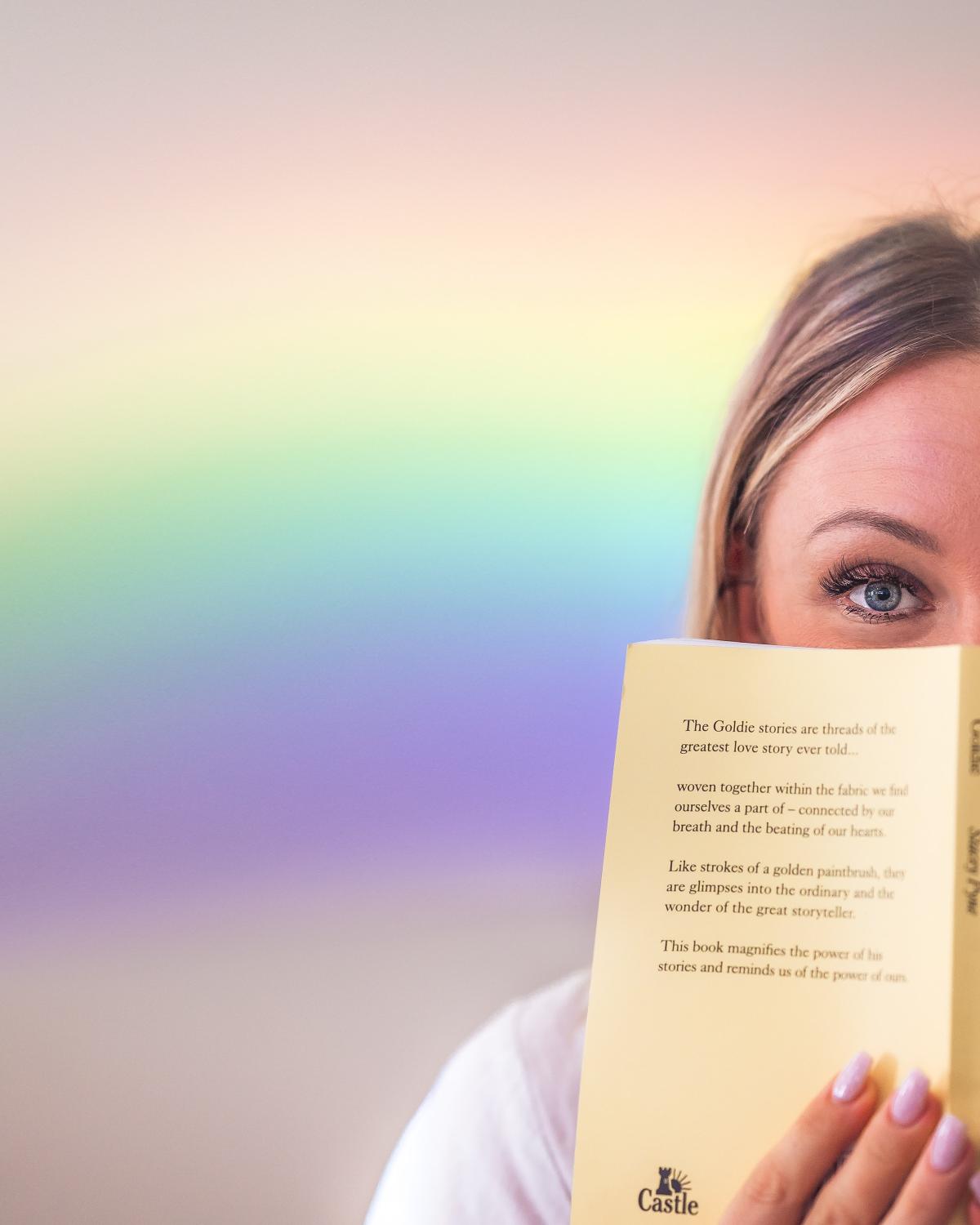 A woman holds a book up in front of her face with a rainbow behind her