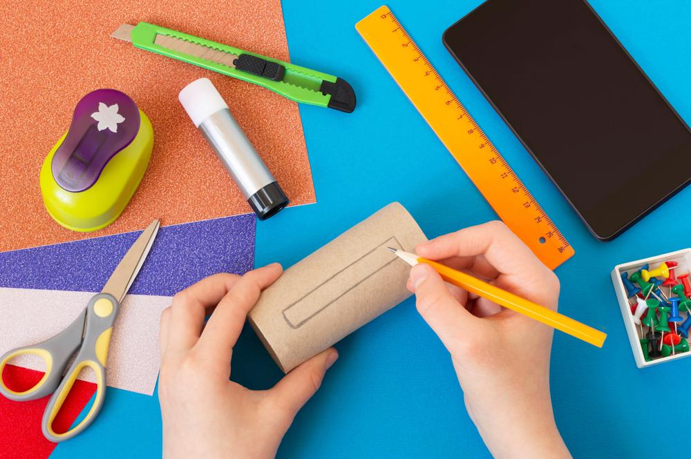 Two hands holding a pencil and a cardboard tube surrounded by various craft supplies