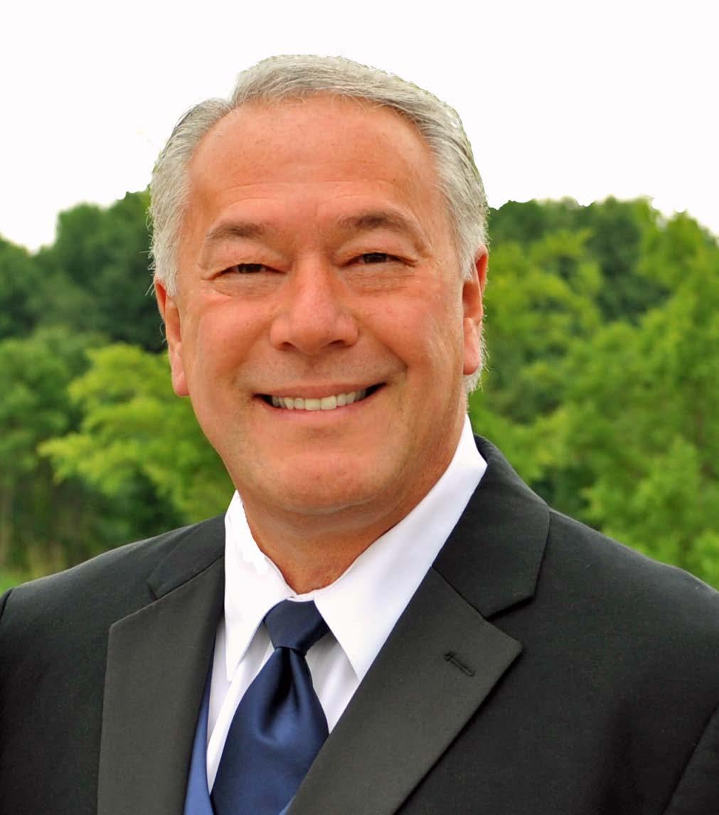 A headshot of a smiling gray haired man wearing a suit jacket and tie in front of green shrubs.