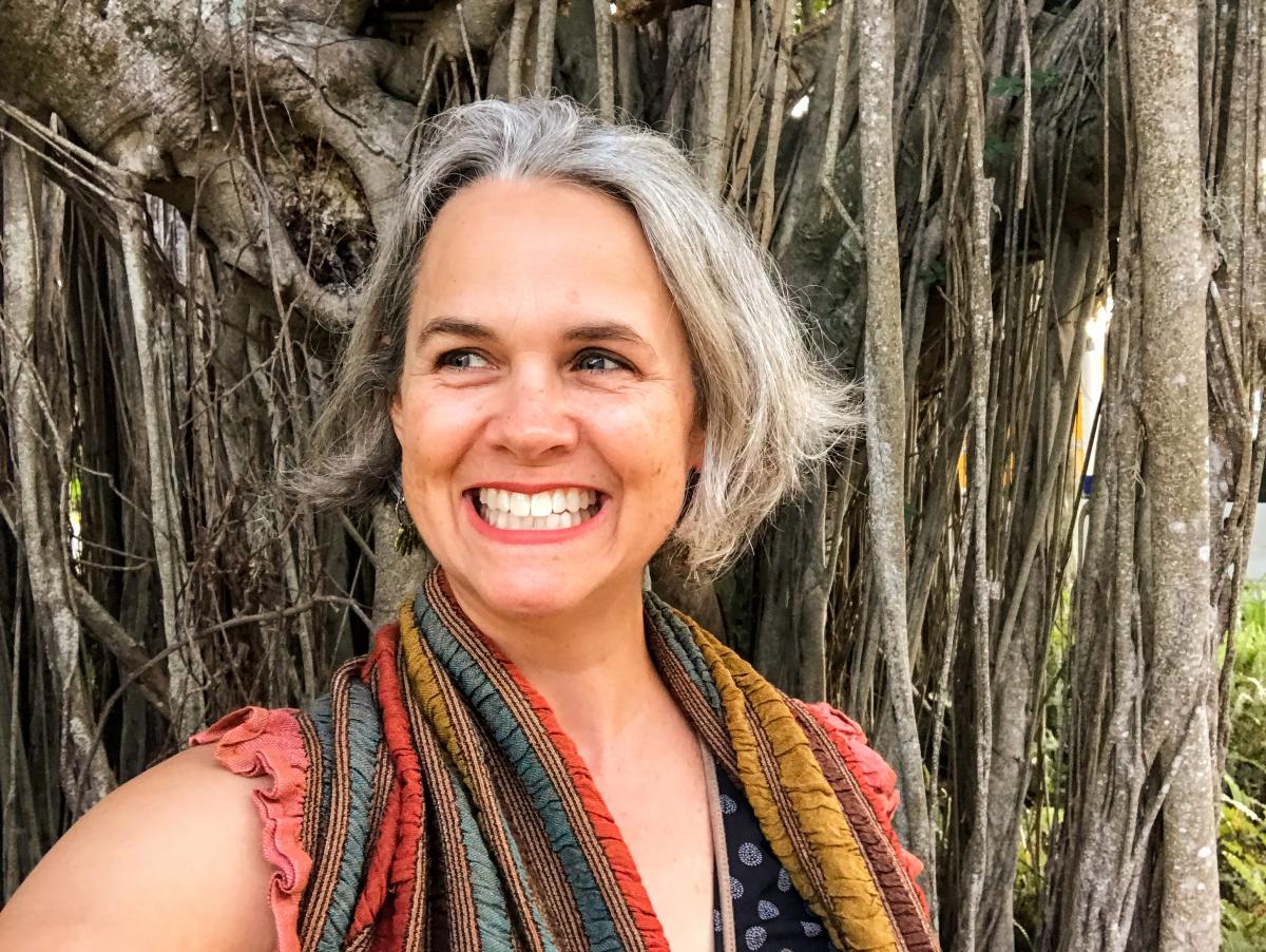 A smiling headshot of poet Laura Shoemaker infront of a banyan tree.