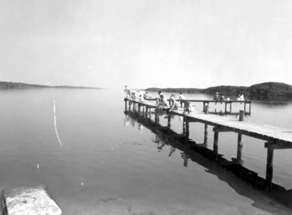 A black and white photograph of people fishing off a pier on a river.