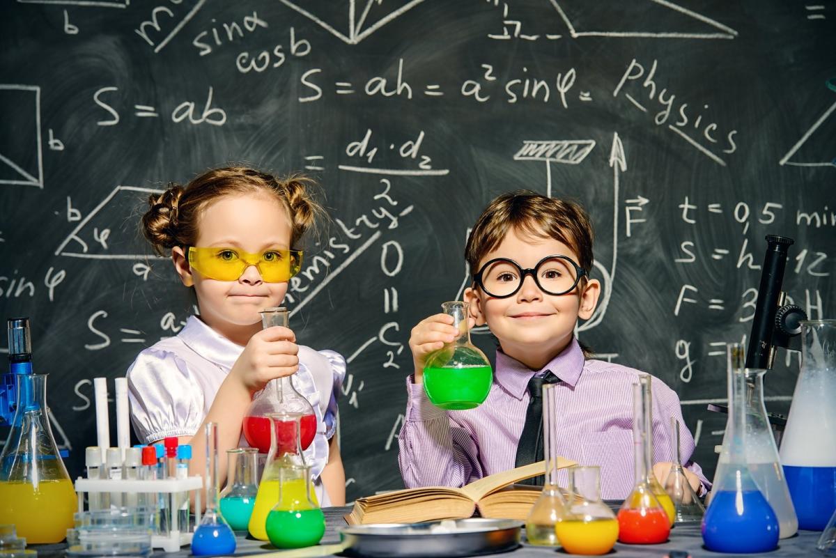 Two children dressed as scientists performing experiments in front of a chalkboard.