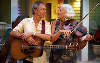 A man holding a guitar and a woman holding a violin, smiling at each other. 